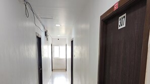 Un long couloir étroit aux murs blancs et au sol carrelé. Des portes se trouvent de part et d’autre, et une enseigne au-dessus d’une de ces portes indique le numéro « 307 ».