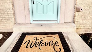 A light blue door with a decorative glass panel, a doormat with "Welcome friends & family" text, and a brick wall to the left. - Luxe 5 BR Family Friendly Oasis-5 King Beds-Hot Tub-Fire Pit-Ski-Hike-Yard Games (Sandy)
