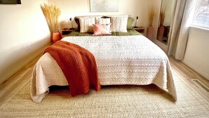 A bedroom with a large bed, a bedside table, a nightstand, a wall-mounted artwork, and a window with curtains. - Luxe 5 BR Family Friendly Oasis-5 King Beds-Hot Tub-Fire Pit-Ski-Hike-Yard Games (Sandy)