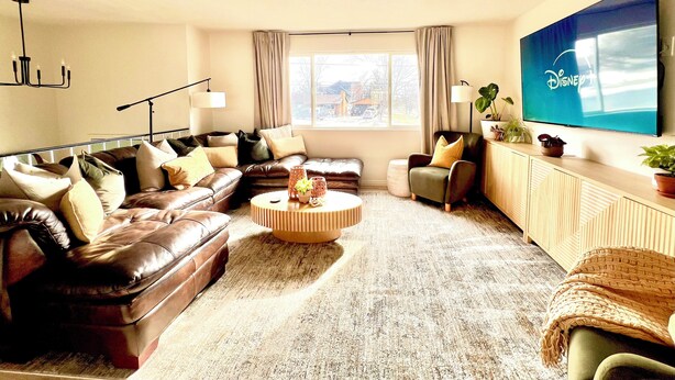 A modern living room with leather sofas, a round coffee table, and a large flat-screen TV mounted on the wall. - Luxe 5 BR Family Friendly Oasis-5 King Beds-Hot Tub-Fire Pit-Ski-Hike-Yard Games (Sandy)
