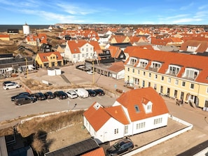 A coastal town with a variety of buildings, including a prominent white building with a red roof, a yellow building, and several other structures with red roofs. There are parked cars in the foreground and a clear view of the sea in the background. - 5 Star Holiday Home in Lokken (Lokken)