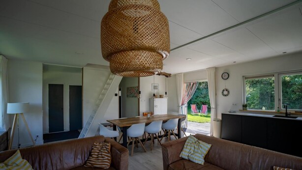 A modern living room with a large dining table, two brown leather sofas, a wicker ceiling light, and a view of the outdoors through large windows. - Holiday Home in Petten With Garden (Petten)