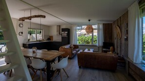 A modern living room with a wooden dining table, white chairs, a brown sofa, and a TV. - Holiday Home in Petten With Garden (Petten)
