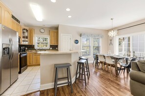 A kitchen with wooden cabinets, a stainless steel refrigerator, a bar stool, a dining area with chairs, and a window with blinds.