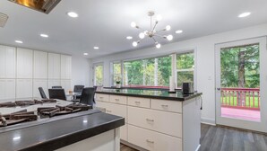 A modern kitchen with white cabinetry, a black countertop, and a large island. There are multiple windows providing natural light and views of greenery outside.