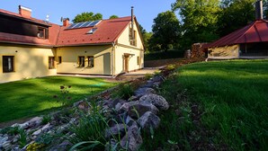 Una casa de dos pisos con techo rojo, una chimenea y un pequeño jardín con muro de piedra y césped.