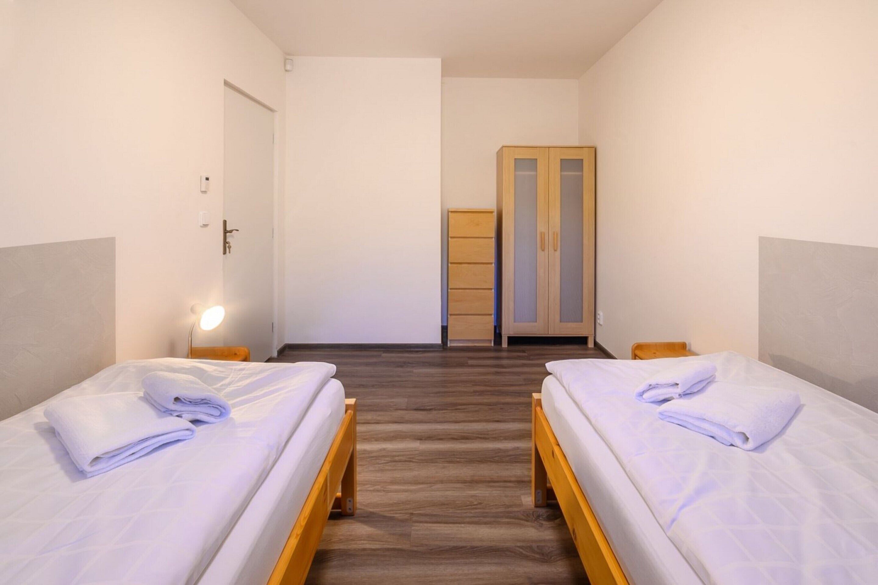 Hotellihuone, jossa on kaksi erillistä sänkyä, puinen vaatekaappi, ovi ja yöpöytä, jossa on lamppu.