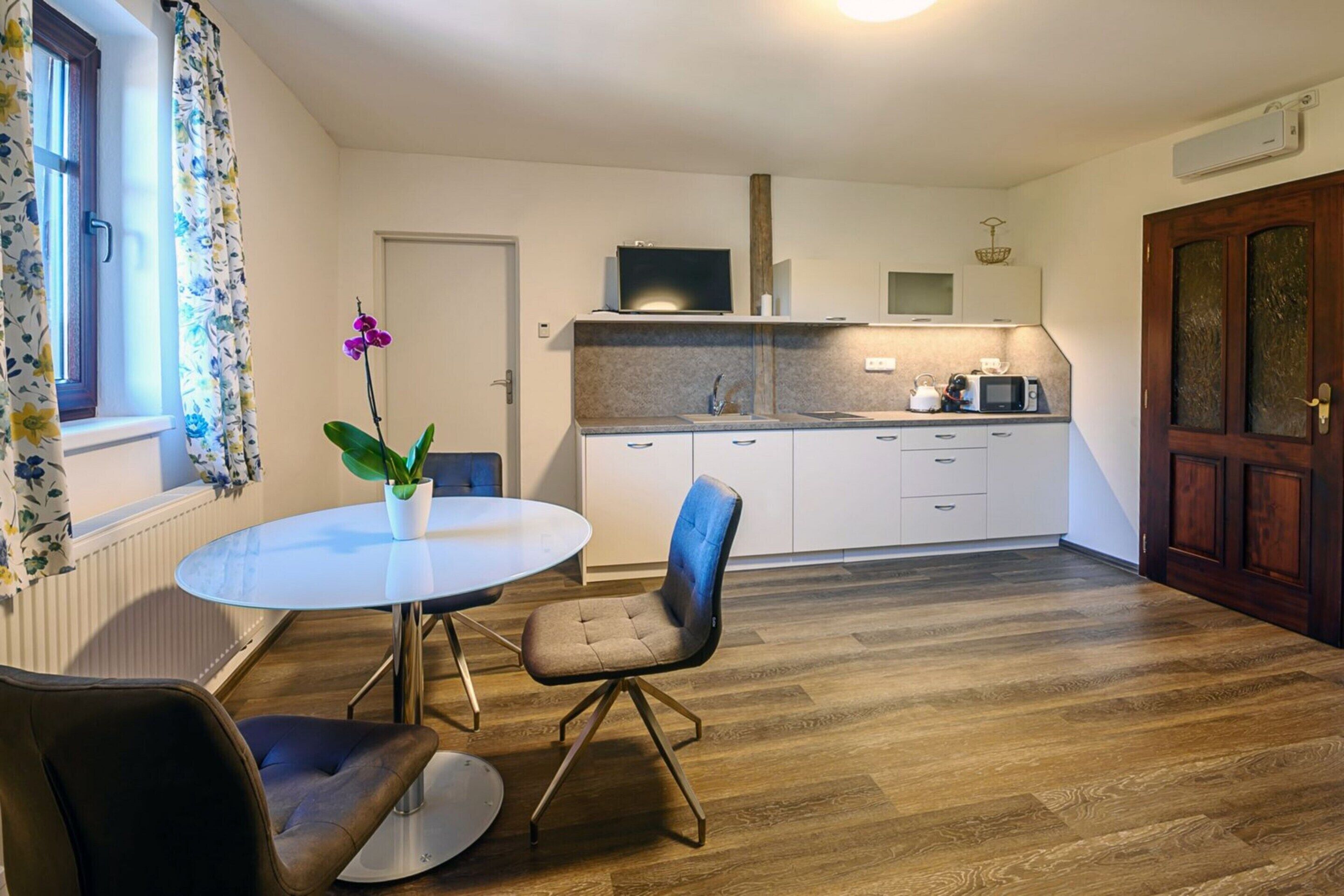 A modern kitchen with white cabinets, a microwave, and a sink. There is a dining table with two chairs and a potted orchid.