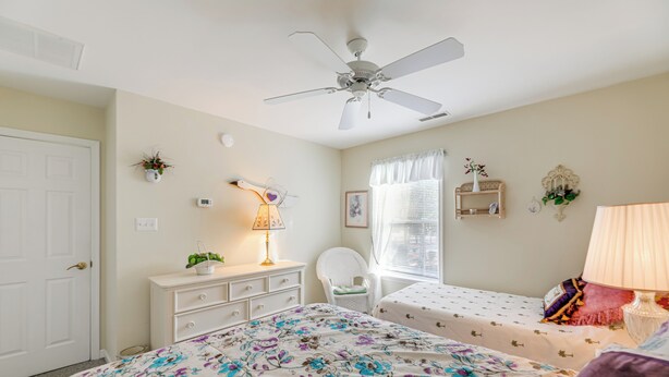 Ein Schlafzimmer mit Bett, Stuhl, Schreibtisch, Deckenventilator und einem Fenster mit Vorhängen.