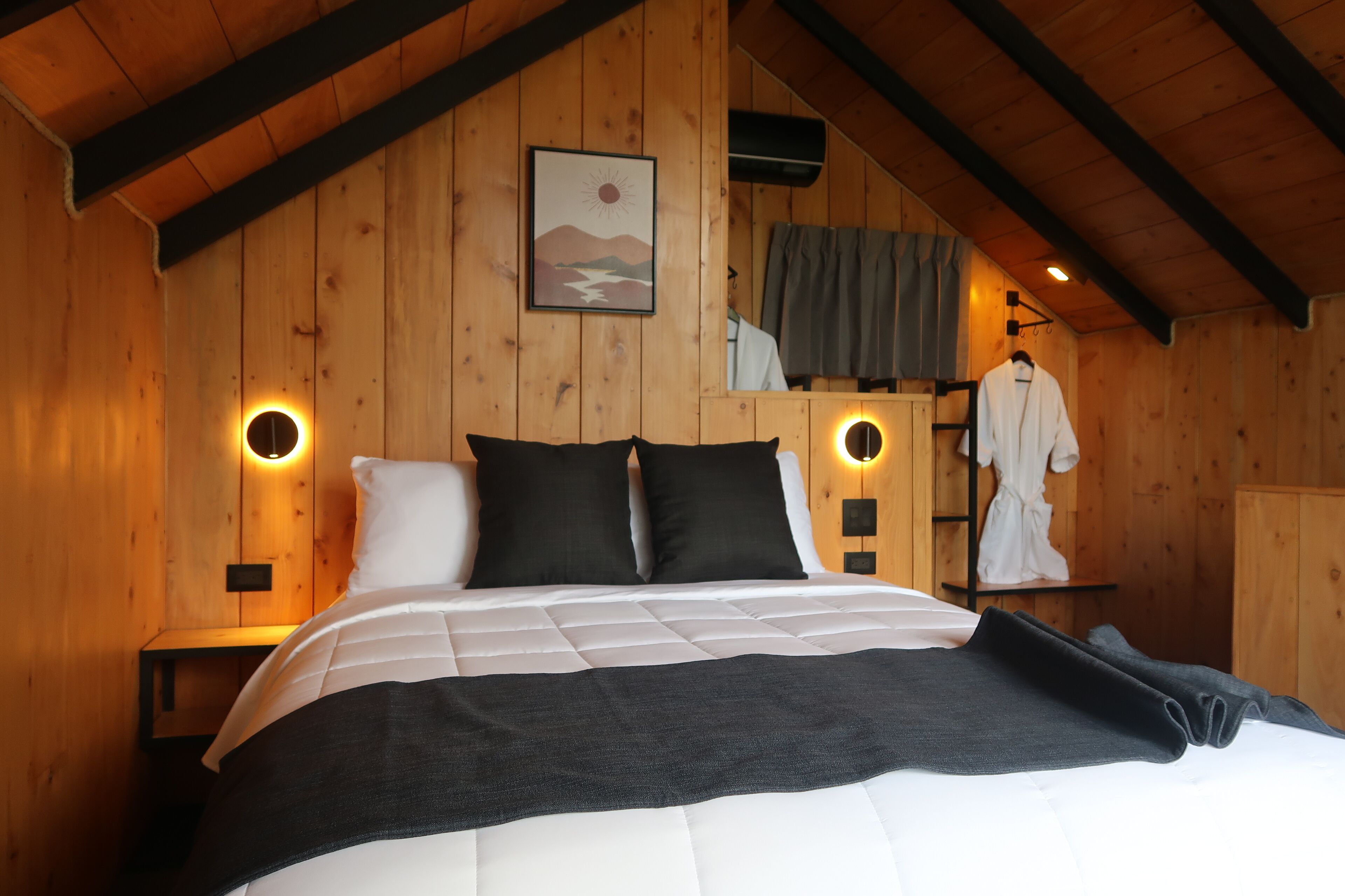 Quarto de cabana de madeira com cama, duas arandelas, um quadro e um roupão pendurado.