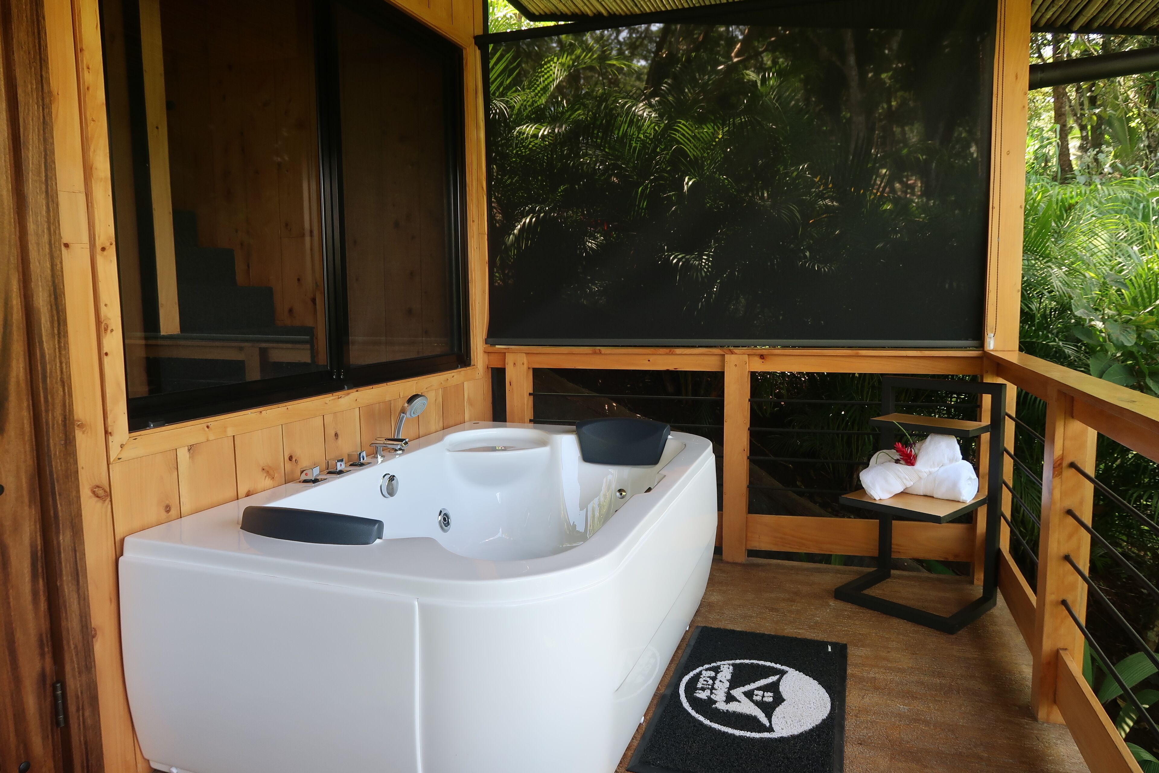 Una cabaña de madera con una bañera blanca, un estante pequeño con toallas y vistas a exuberante vegetación.