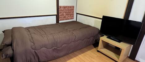 Uma cama de solteiro com edredom marrom, uma televisão sobre suporte de madeira e uma parede de tijolos decorativa.