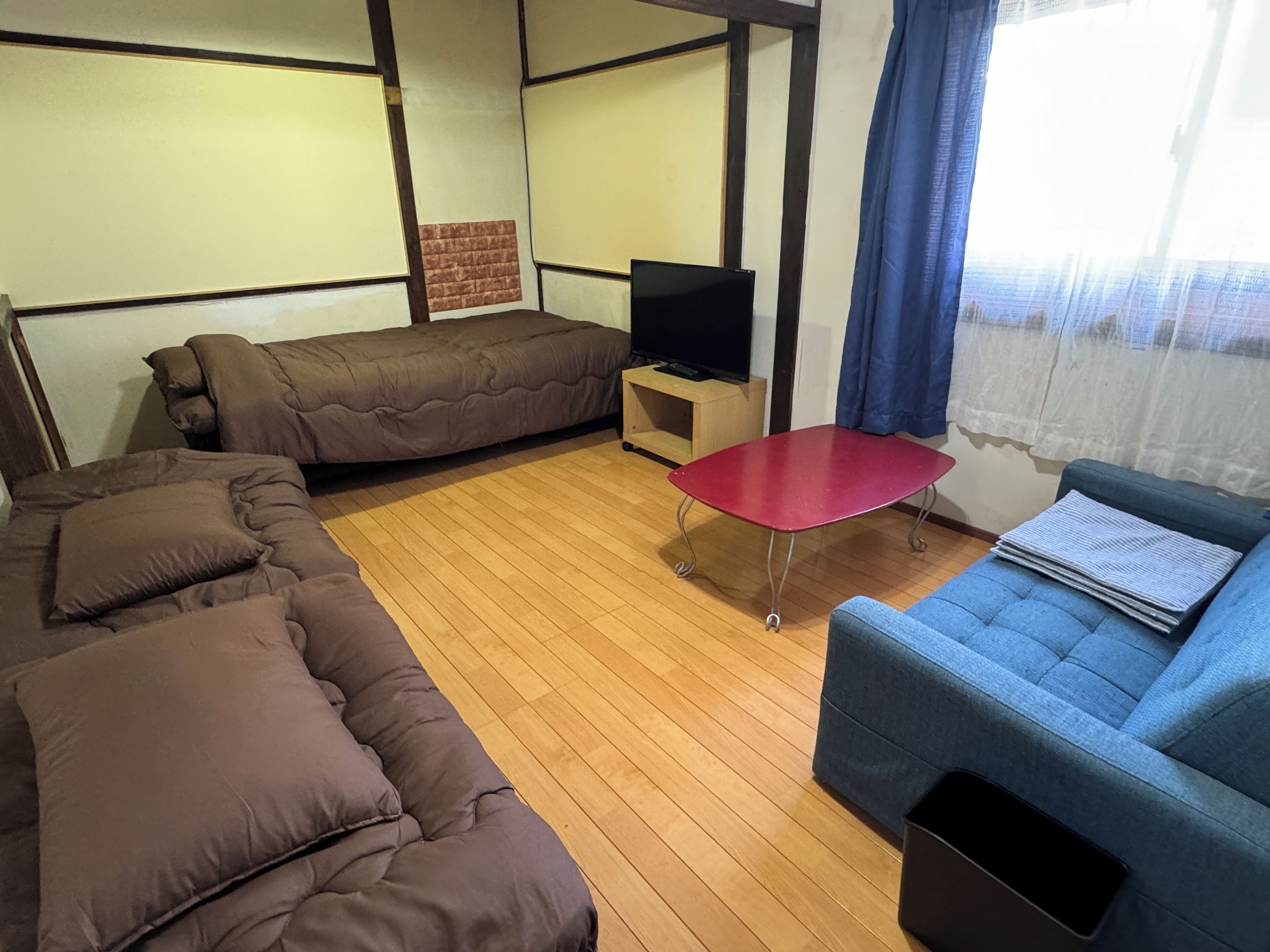 Una habitación con un sofá seccional, un sillón azul, una mesa roja y un televisor sobre un mueble de madera.