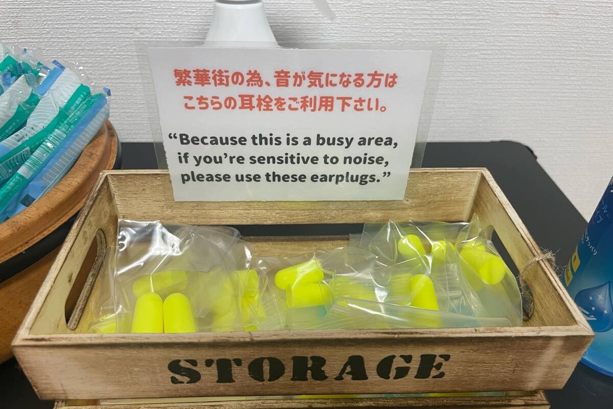 A wooden storage box labeled 'STORAGE' with the number '02' on it, containing yellow earplugs and a sign advising noise-sensitive individuals to use them.