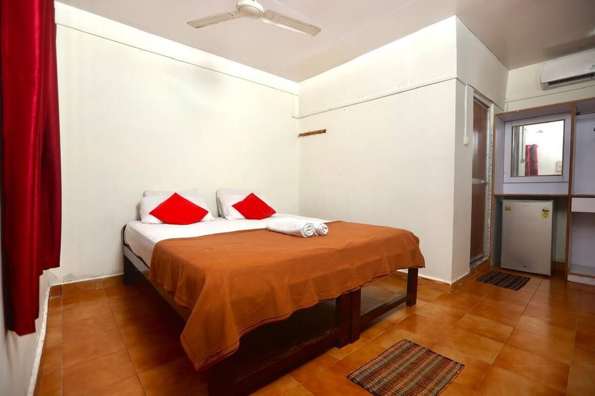 Un lit simple avec une couvre-lit marron, des oreillers blancs et quelques oreillers décoratifs rouges. On y trouve également un ventilateur de plafond, un petit réfrigérateur et une porte dotée d’un miroir.