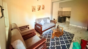 A living room with a sofa, two wooden chairs, a glass coffee table, and a patterned rug. There are three framed pictures on the wall and a ceiling light. - The Village Harmony by Red Rose (Canacona)