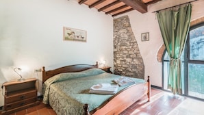 A bedroom with a wooden bed, a stone wall, a window with curtains, and a nightstand with a lamp.