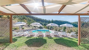 A pool area with sun umbrellas and lounge chairs, surrounded by a wooden pergola and a view of a hilly landscape.