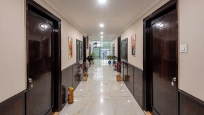 A long, narrow hallway with dark wooden doors on either side, potted plants along the walls, and a fire extinguisher on the floor. - DLS Park Grace (Haridwar)