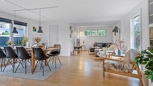 Un vaste espace de vie comprenant une table à manger, des chaises et un canapé. De grandes fenêtres laissent entrer la lumière naturelle et offrent une vue sur la verdure environnante.