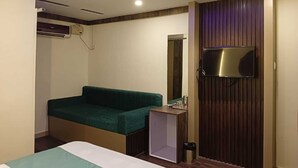 A hotel room with a bed, a green sofa, a small table, a TV mounted on a wooden panel, and a ceiling fan. - Seven Seas Dream Castle Port Blair (Port Blair)