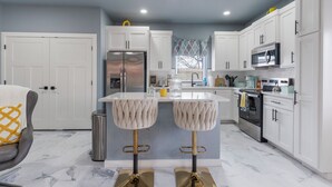 A modern kitchen with white cabinets, a stainless steel refrigerator, a microwave, and a central island with bar stools. - Madison's Masterpiece in Downtown Tulsa-Sleeps 6 (Tulsa)