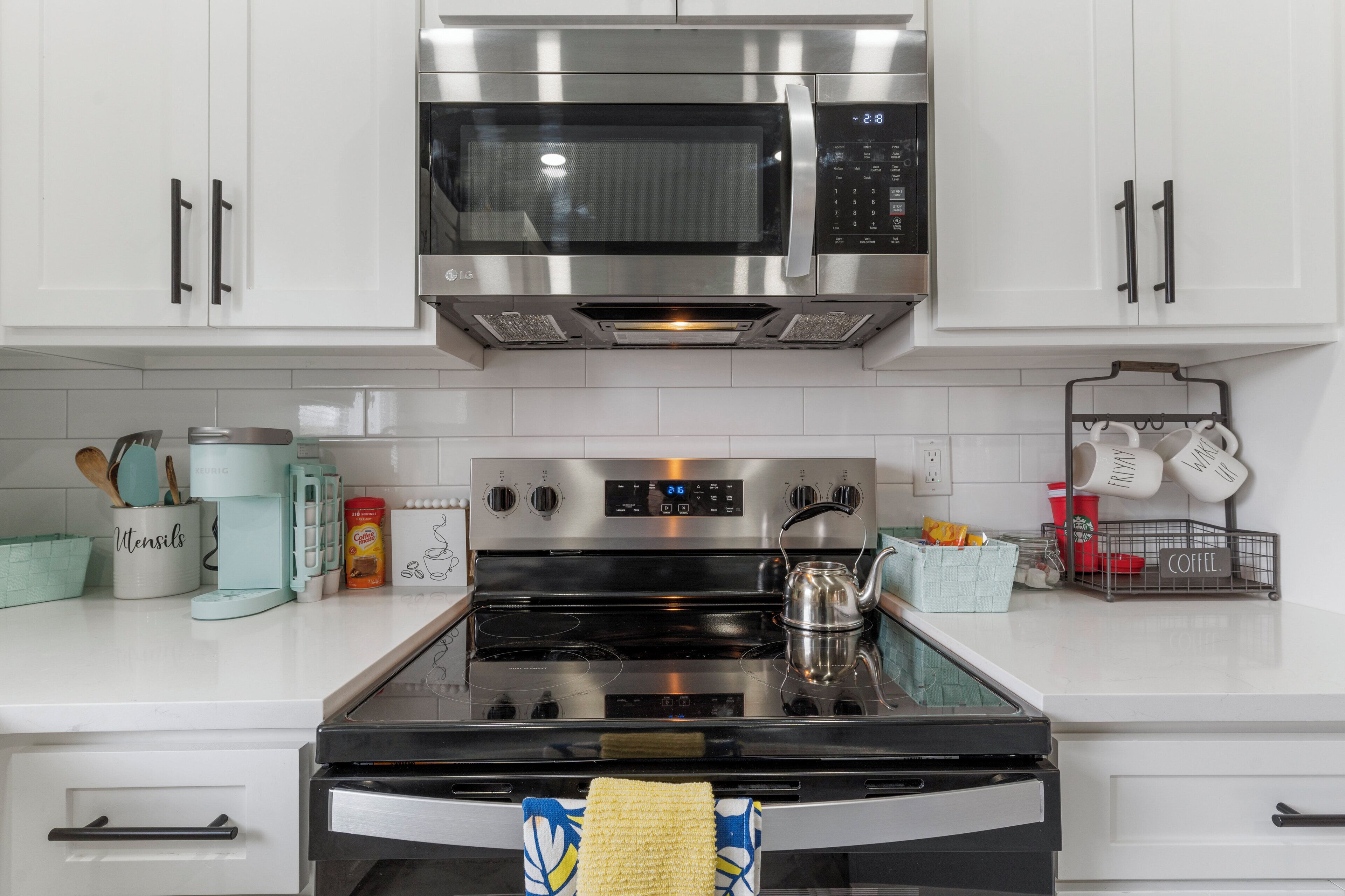 Una cocina con gabinetes blancos, un microondas de acero inoxidable, una estufa y diversos utensilios y electrodomésticos.
