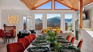 A dining area with a long table set for a meal, large windows with a mountain view, and a neon sign reading "LAKE LURE BAR".