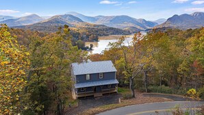 A house with a sloped roof, surrounded by trees and a lake, with mountains in the background.