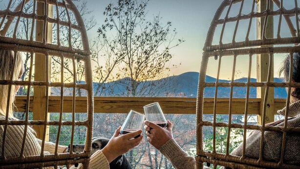 Two people sitting in wicker chairs with a view of trees and mountains through a wooden railing.