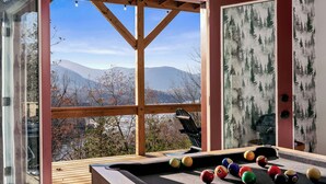 A pool table with balls, a wooden deck, and a view of mountains through a window.