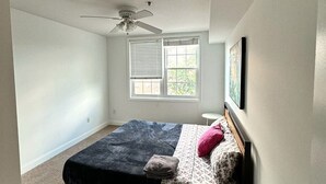 Ein Schlafzimmer mit einem Bett, einem kleinen runden Tisch, einem Deckenventilator, einem Fenster mit Jalousien und einem gerahmten Bild an der Wand.