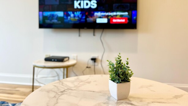 Ein an der Wand befestigter Fernseher zeigt verschiedene Sender, ein kleiner Tisch mit einer Fernbedienung und ein Blumentopf auf einem Marmortisch.