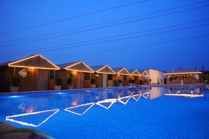 Una piscina con patrones de luz geométricos, edificios de madera con techos triangulares y un cielo azul despejado.