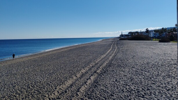 Une plage où l’on voit une personne se promener, un ciel dégagé et des bâtiments en arrière-plan.