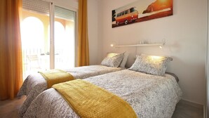 Une chambre avec deux lits, une fenêtre avec des rideaux, un tableau représentant un fourgon et une étagère avec divers objets.