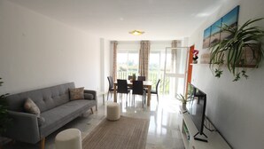 Un salon moderne avec un canapé gris, un petit pouf, un téléviseur à écran plat et un coin repas comprenant une table et des chaises.