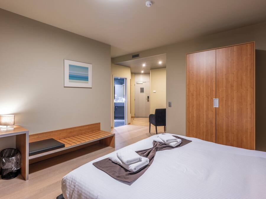 Une chambre d’hôtel avec un lit, un bureau, une chaise, une armoire en bois et un petit banc.