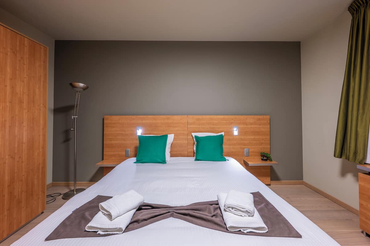Une chambre d’hôtel moderne avec un grand lit, deux oreillers verts, une tête de lit en bois, une table de chevet avec une plante et une lampe sur pied.