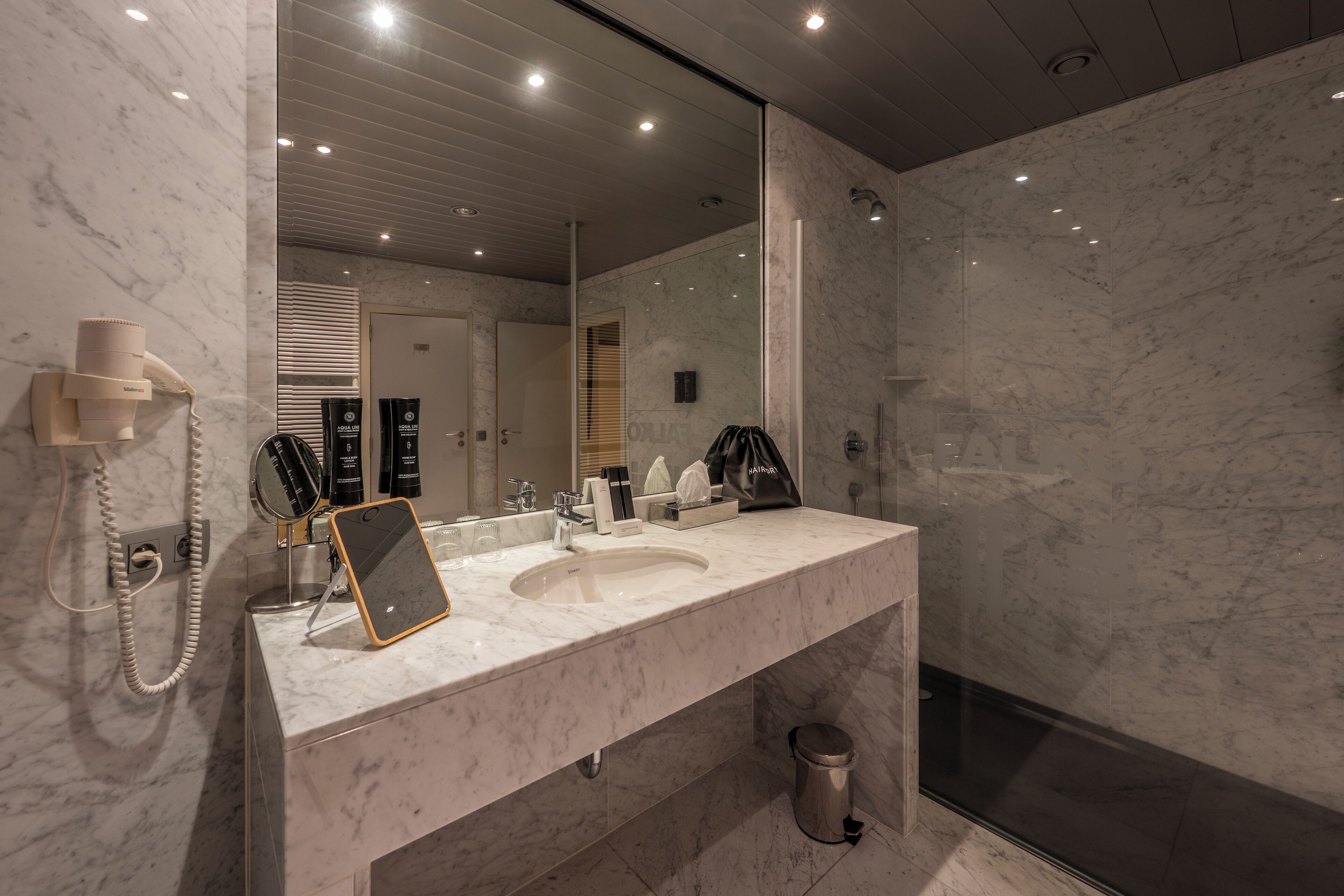 A modern bathroom with a marble sink, a large mirror, a handheld showerhead, and a hairdryer.