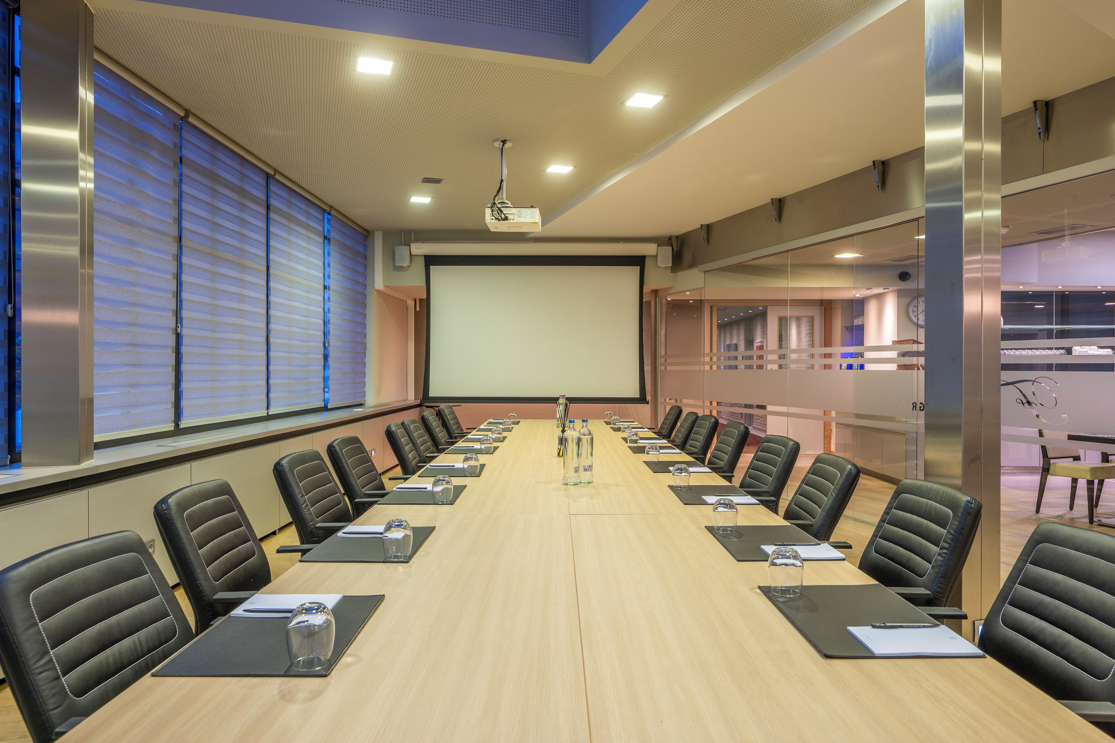 Un salón de conferencias con una larga mesa, sillas de cuero negro, una pantalla de proyector y un amplio ventanal con persianas.