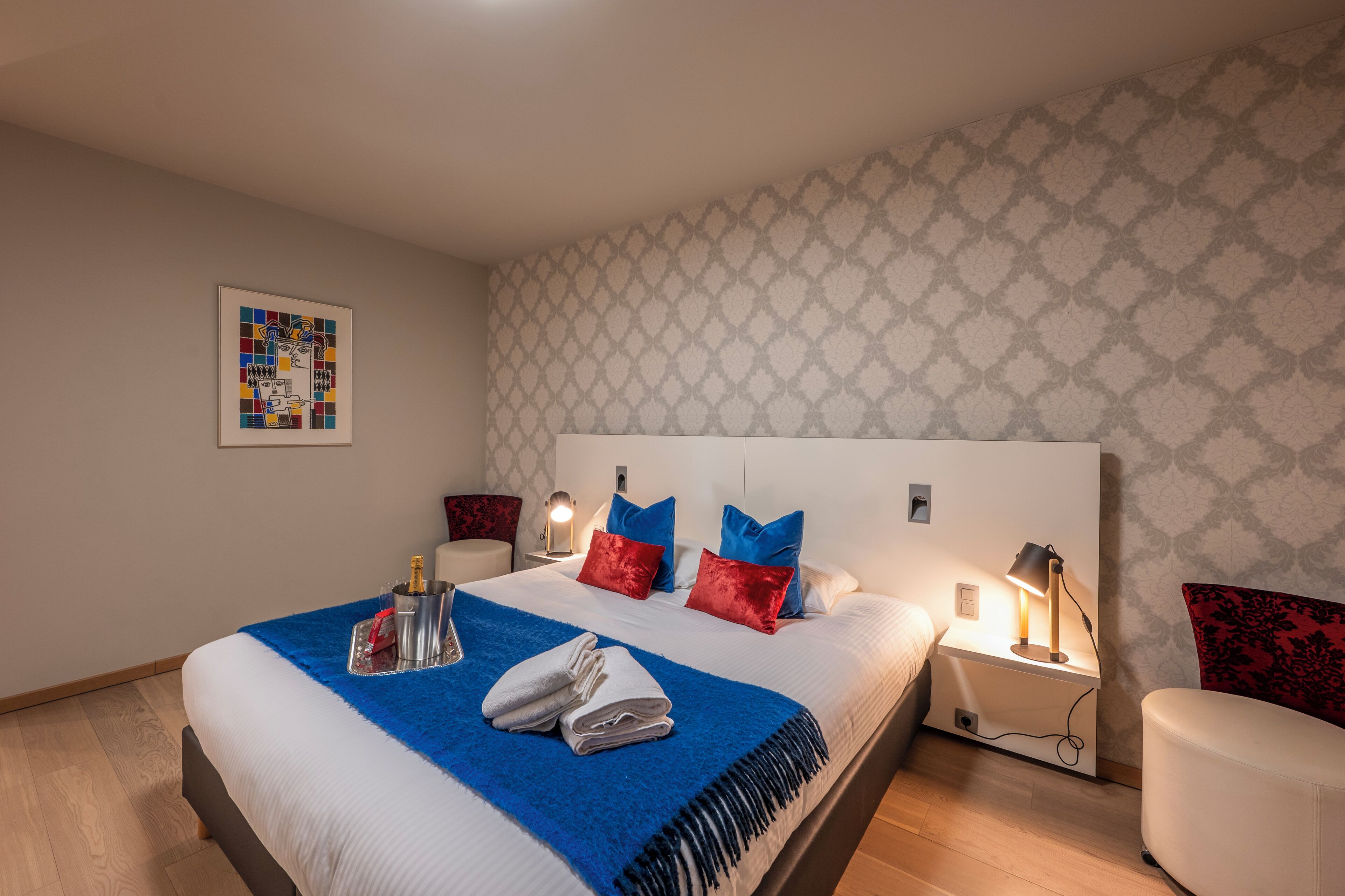 Hotelkamer met een groot bed, een blauwe plaid en rode kussens. Er staat een nachtkastje met een lamp en een fles champagne. De kamer heeft behang met een patroon en een ingelijst schilderij aan de muur.