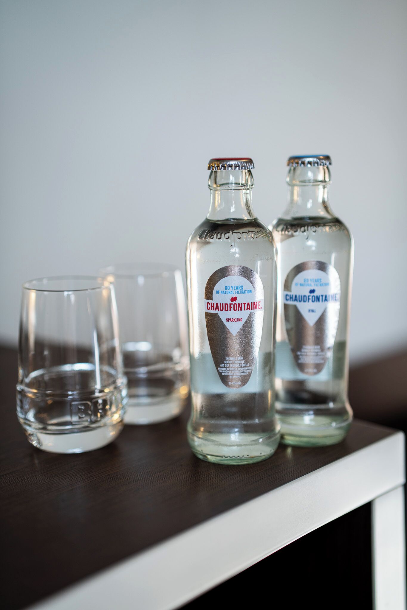 Two bottles of Chaudfontaine sparkling water and two empty glasses on a dark wooden surface.
