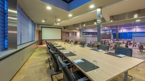 A conference room with a long table, chairs, a projector screen, and a large window with blinds. - Falko Hotel (Meise)