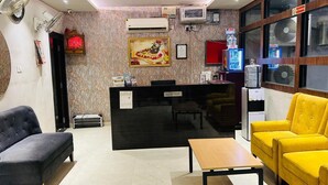 A reception area with a reception desk, a sofa, a yellow armchair, a small table, and a wall-mounted air conditioner. - Hotel Aalishan (Varanasi)