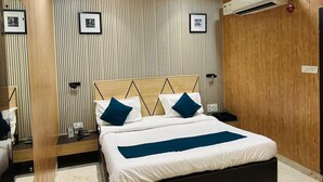 A modern bedroom with a wooden headboard, a bed with white and blue bedding, a nightstand with a lamp, and an air conditioning unit on the wall. - Hotel Aalishan (Varanasi)