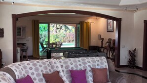 A living room with a sofa, a coffee table, and a pool table. There is a window with curtains, a door leading to a balcony, and a ceiling fan. - Paparadise, a haven of peace in the heart of Papara on the island of Tahiti. (Paparā)
