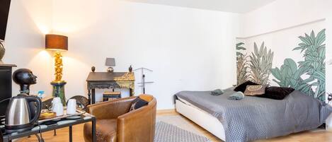 Un dormitorio con cama, una silla, una mesa con hervidor, una lámpara y un mural de plantas en la pared.
