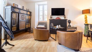 A living room with a wooden floor, a dark wooden cabinet with decorative panels, a brown leather sofa, a small table with a lamp, and a television mounted on the wall. - L'Eclose - Kergoz, room in a private home in Guilvinec (Guilvinec)