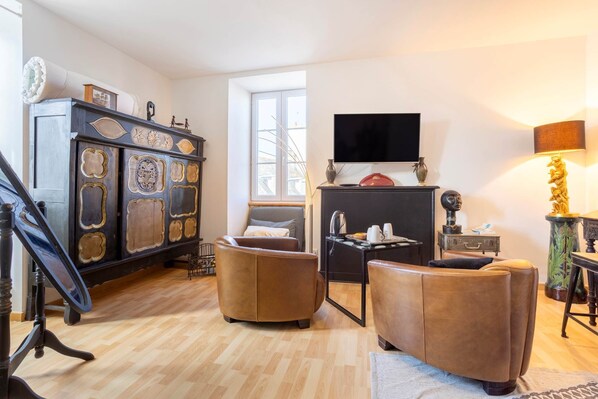 A living room with a wooden floor, a dark wooden cabinet with decorative panels, a brown leather sofa, a small table with a lamp, and a television mounted on the wall.